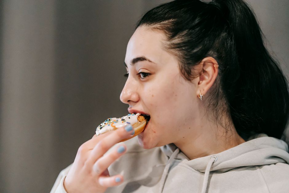 Crop young brunette in comfy hoodie eating fresh appetizing doughnut covered with vanilla icing and sprinkles in light room