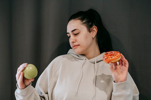 Pensive female wearing casual hoodie deciding between healthy green apple and sweet doughnut in studio