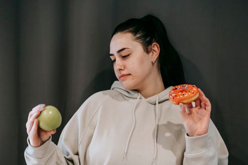 Free Pensive female wearing casual hoodie deciding between healthy green apple and sweet doughnut in studio Stock Photo