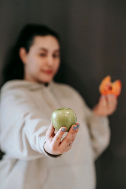 Blurred female in casual wear choosing between ripe green apple and sweet donut and showing apple to camera