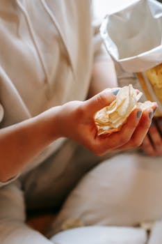 From above crop anonymous female in comfy wear demonstrating savory potato chips while sitting on floor