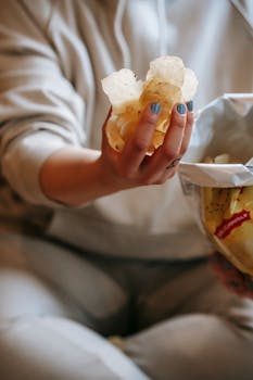 A close-up image of a female hand holding crispy potato chips in a relaxed indoor setting.