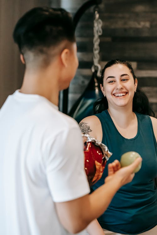 Free Anonymous young man offering apple and potato chips to cheerful obese female friend in gym Stock Photo