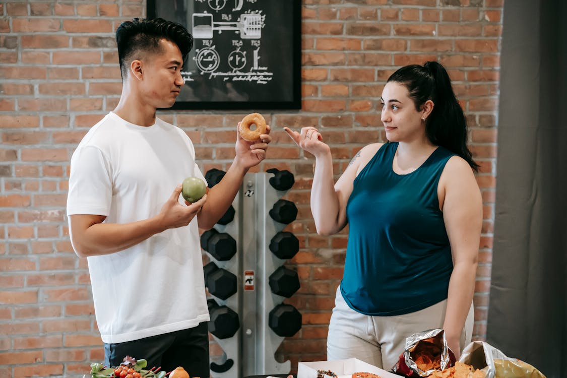 Side view of Asian personal instructor with apple and doughnut offering healthy food to plus size woman while standing in gym