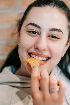 Smiling woman savoring a delicious chip with joy indoors, against a brick wall.