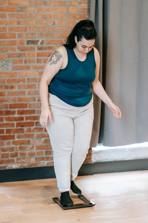 Full body of plus size female in activewear standing on weighing scales near brick wall in spacious gym after training