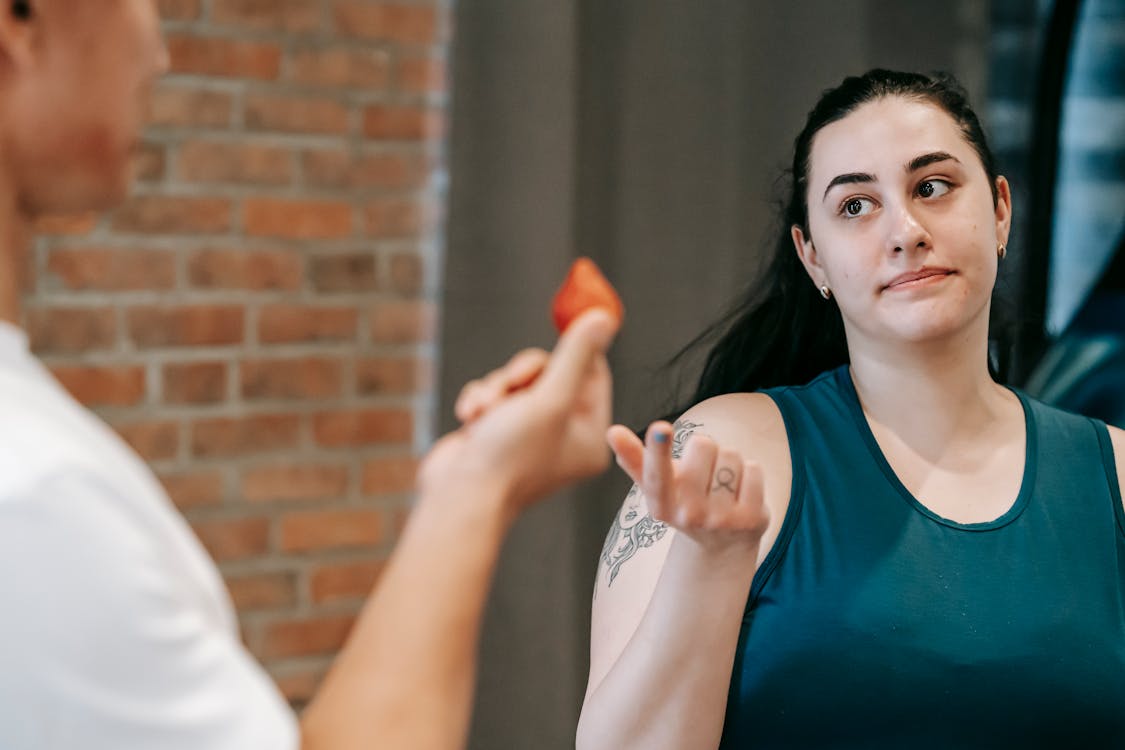 A woman in workout attire gestures as a man offers her a strawberry in a gym setting.