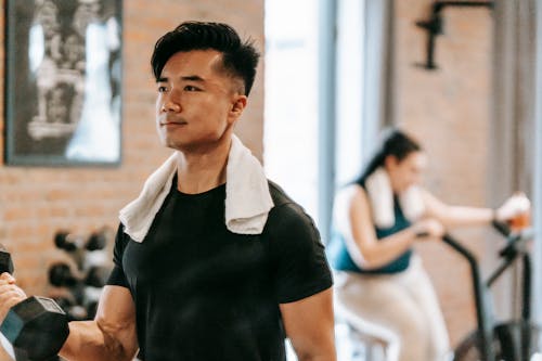 Free Determined Asian male athlete with towel lifting heavy dumbbell while training in spacious gym with plump woman on blurred background Stock Photo