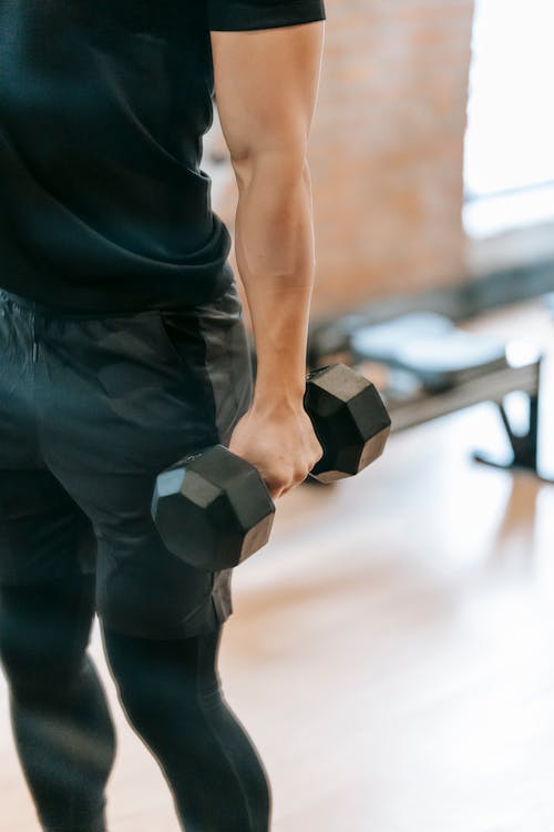 Unrecognizable muscular male in black sportswear lifting heavy dumbbell during weightlifting workout in light gym with bench on blurred background