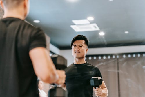 Strong Asian male in activewear training biceps with heavy dumbbells while looking in reflection of mirror during workout in modern fitness studio