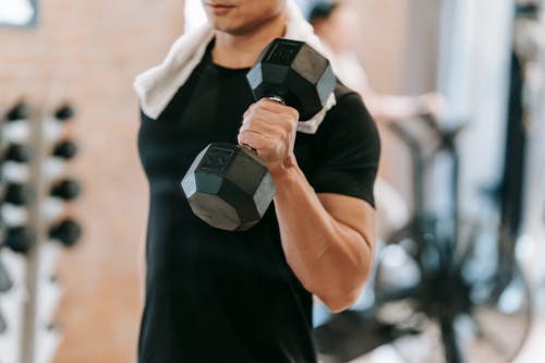 Free Unrecognizable strong male with towel wearing black activewear lifting heavy dumbbell during intense training in fitness studio with special equipment on blurred background Stock Photo