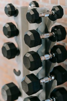 Stack of black heavy dumbbells for weightlifting placed on metal stand near brick wall in modern fitness studio during workout