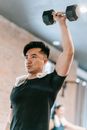 Sportive Asian male in black sportswear with raised arm lifting metal dumbbell while training in fitness studio with woman on blurred background