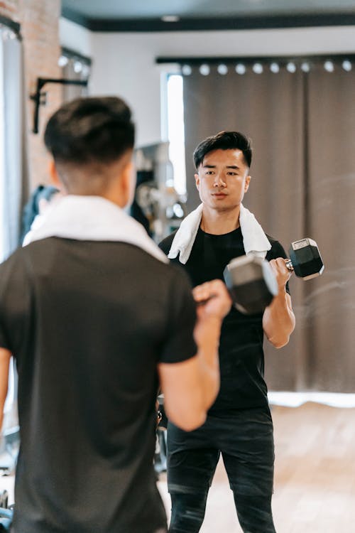 Determined Asian male in activewear with towel doing dumbbell curls while standing near mirror during fitness training in modern spacious gym