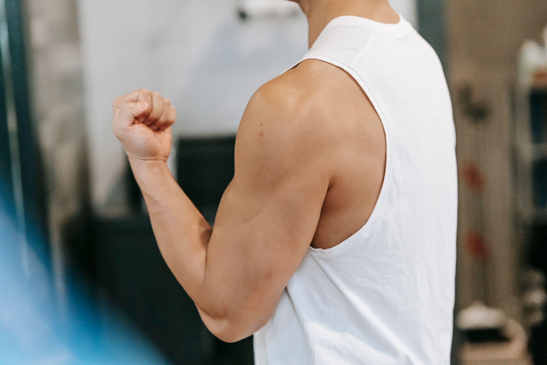 Side view of unrecognizable muscular male in white sportswear standing with bent arm while showing bicep during workout in gym on blurred background