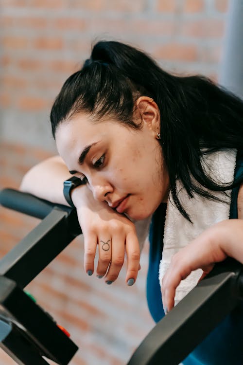Free Side view of tired ethnic female with towel looking down while standing near modern fitness equipment during workout on blurred background Stock Photo
