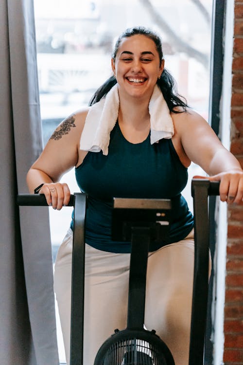 Smiling plus size female with towel exercising on cross trainer machine while looking at camera during workout in fitness studio