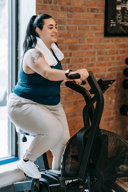 Free Side view of cheerful plus size ethnic female in activewear with towel exercising on fitness equipment during workout in gym Stock Photo