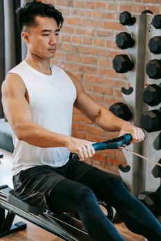 From above of young concentrated ethnic male working out on rowing machine in sport club with brick walls