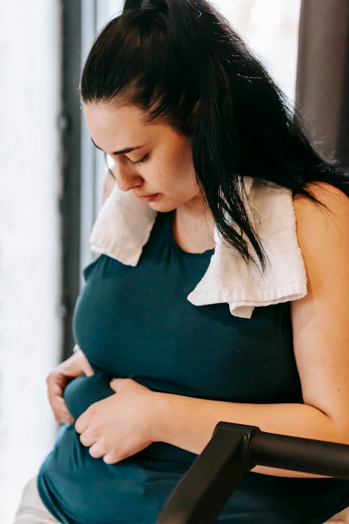 Crop young plus size female with towel on shoulders showing big belly in gym