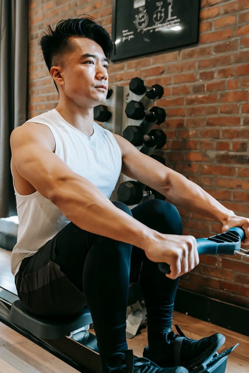 Young ethnic male in sportswear training on rowing machine while looking forward in sport club with brick walls