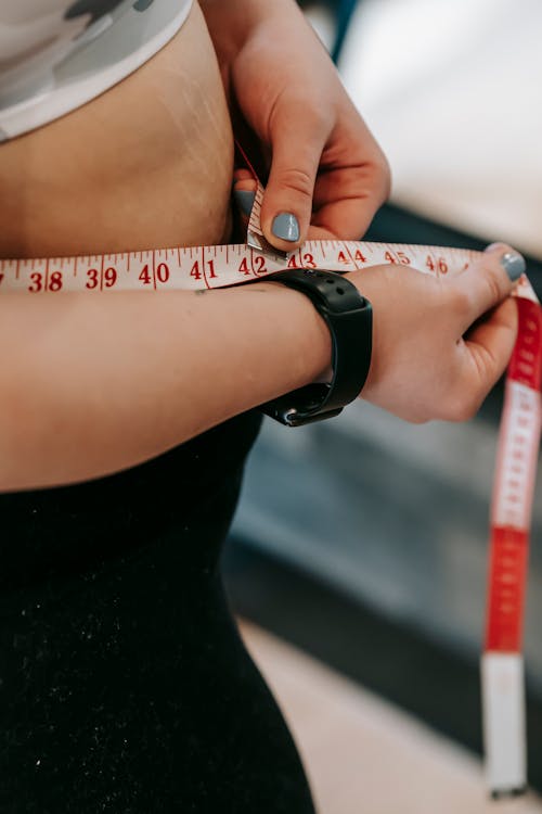 Crop anonymous female with measuring tape around big belly on blurred background of gym