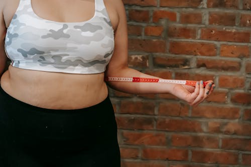 Crop anonymous plump female using measuring tape for measuring belly near brick wall