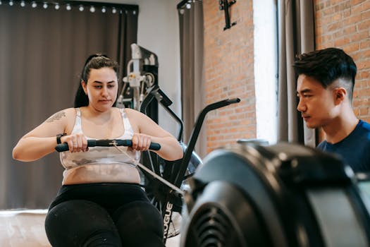 A woman works out on a rowing machine with guidance from a personal trainer in a gym.