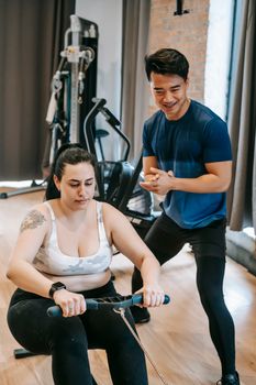 From above of positive ethnic trainer smiling and helping plus size female doing exercise on rowing machine in gym