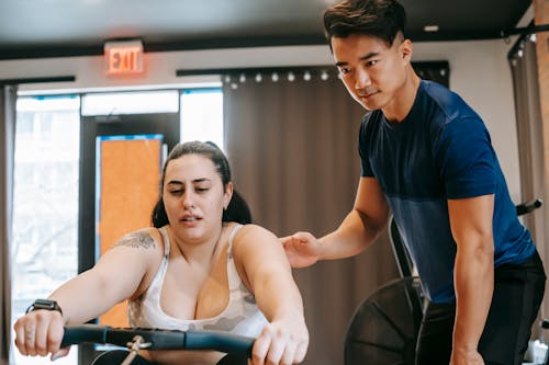 Free Young handsome ethnic experienced trainer supporting plus size woman working out on rowing machine in gym Stock Photo