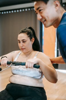 Concentrated plus size female with long dark hair doing weigh lose exercise on rowing machine while exercising in modern gym with crop positive Asian male trainer
