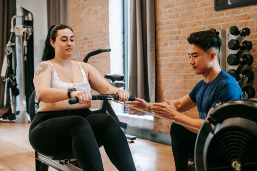 Positive young obese tattooed female in activewear sitting on rowing machine and doing pulling exercise during intense training with Asian male personal coach in modern gym