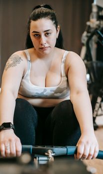 Thoughtful young plus size lady with long dark hair in sportswear doing weight lose exercise on rowing machine and looking at camera in modern gym