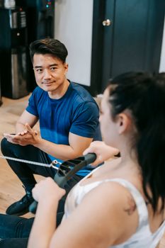 From above of positive young Asian male trainer looking at determined overweight lady doing pulling exercise on gym equipment during intense workout