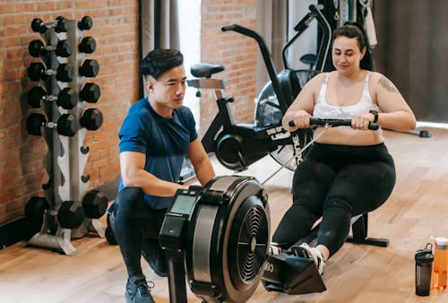 Positive young plus size female in leggings and crop top doing abs exercise on rowing machine during intense workout in modern gym with fit Asian male trainer