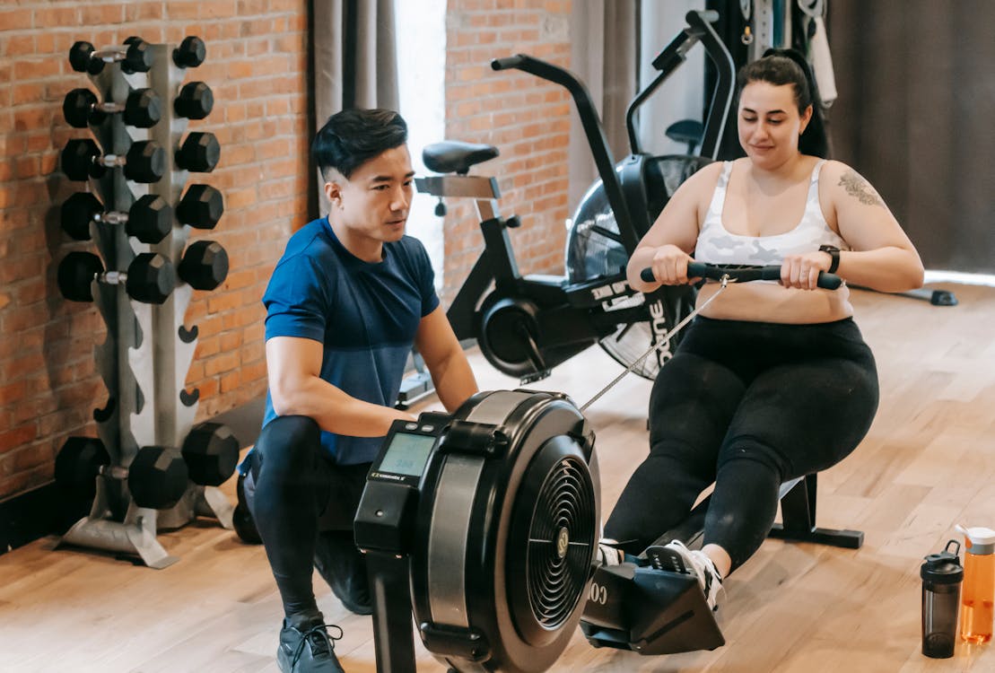 Free Positive young plus size female in leggings and crop top doing abs exercise on rowing machine during intense workout in modern gym with fit Asian male trainer Stock Photo