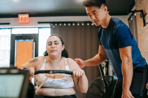 Determined young overweight female exercising on gym machine during training with ethnic coach