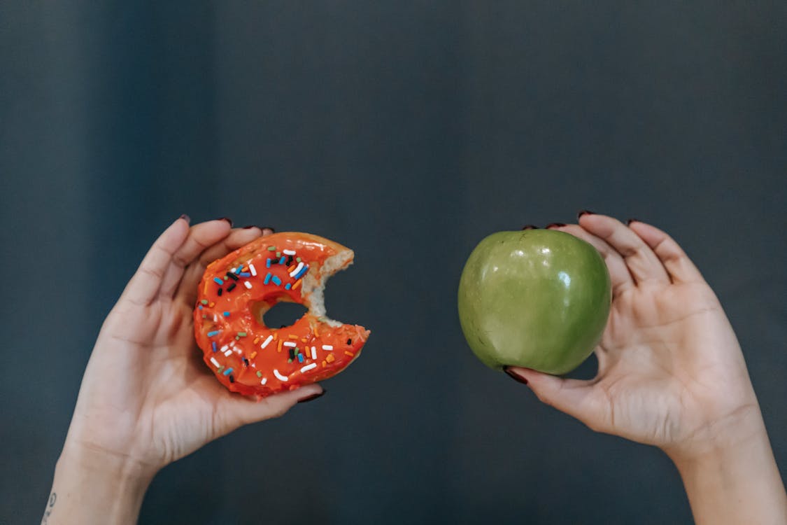Free Crop unrecognizable young female demonstrating bitten high calorie doughnut and healthy green apple against gray background Stock Photo