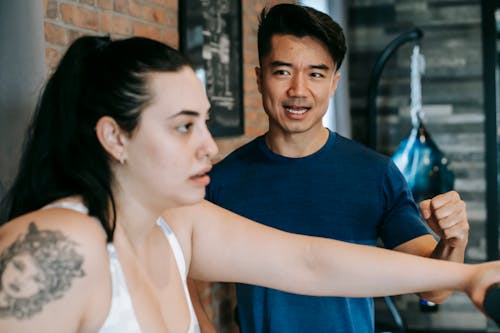 Side view of serious young exhausted obese woman exercising on gym equipment near determined Asian male instructor