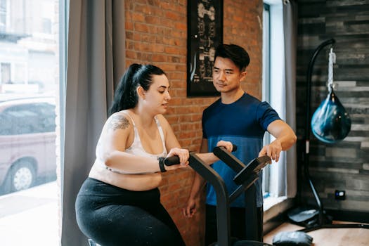 Young concentrated plus size lady in sportswear training on cycling machine during cardio workout in modern fitness club with ethnic male personal trainer