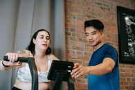 Young Asian male trainer checking fitness equipment while instructing overweight female