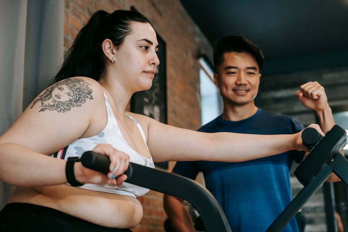 Curvy woman From below of happy young Asian male trainer motivating exhausted young curvy female client to to cardio fat burning exercise on elliptical machine in modern sport club Stock Photo