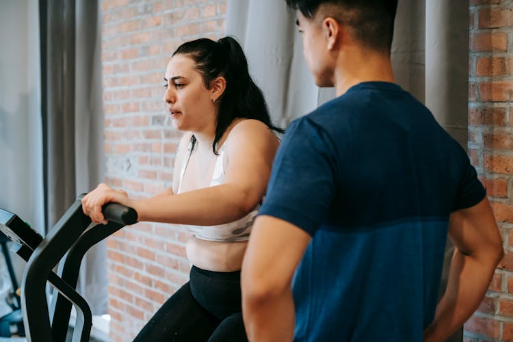 Serious Young Asian Male Trainer Looking At Overweight Woman Exercising On Gym Bike