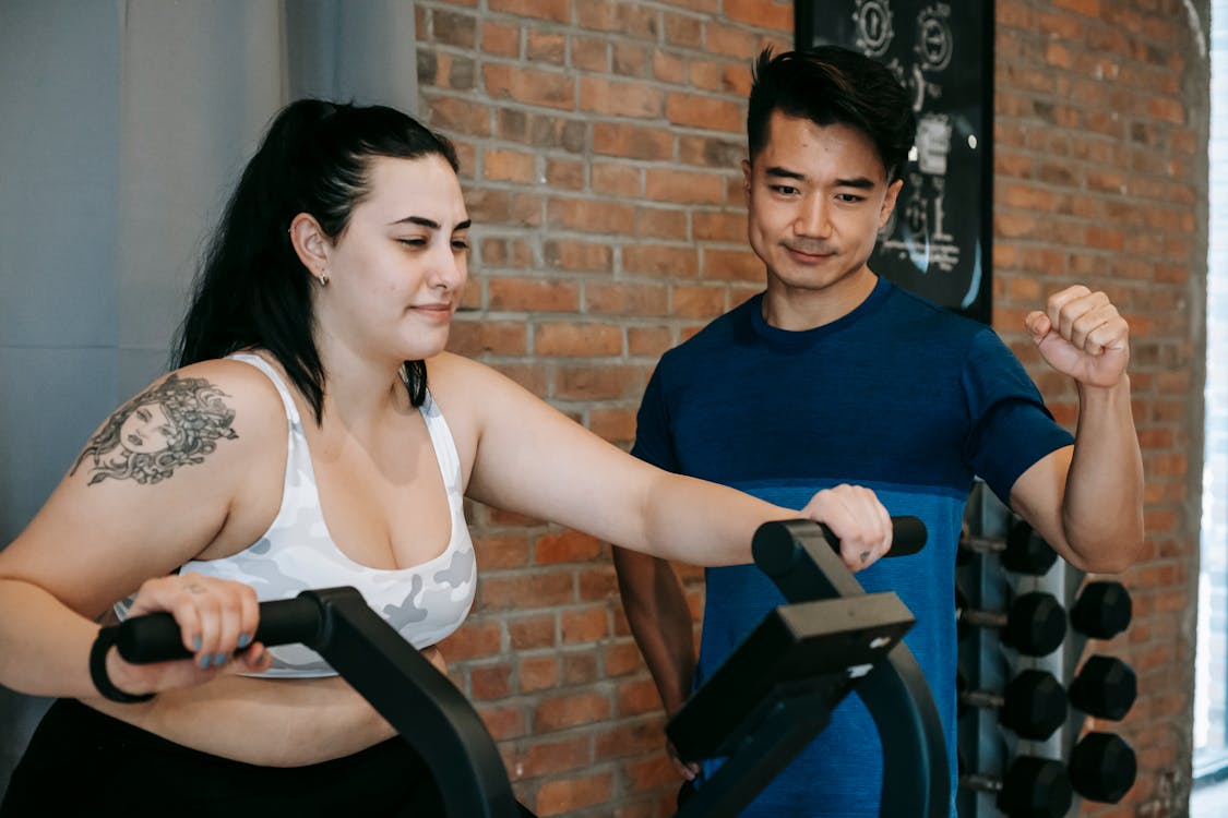 Free Smiling young tattooed plus size sportswoman in activewear doing cardio exercise on fitness cycle while training in contemporary gym with positive Asian male instructor Stock Photo