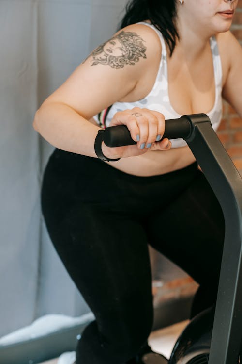Side view of crop unrecognizable tattooed plus size lady in activewear exercising on cycling equipment during weigh lose workout in gym