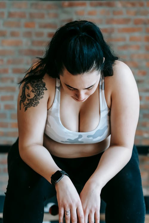 Exhausted young tattooed overweight female with ponytail in sportswear and smart watch sitting on bench in gym and resting after hard workout