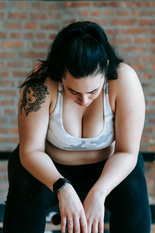 Free Exhausted young tattooed overweight female with ponytail in sportswear and smart watch sitting on bench in gym and resting after hard workout Stock Photo