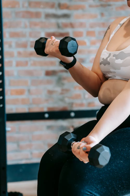 Side view of crop anonymous plus size woman in activewear lifting dumbbells during weight lose training in modern sports center