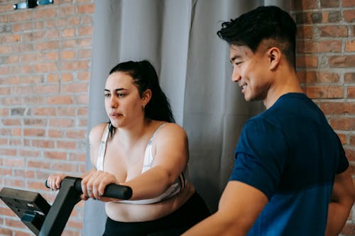 Side view of positive young Asian male trainer supporting plus size female doing cardio workout on cycling machine in modern fitness center