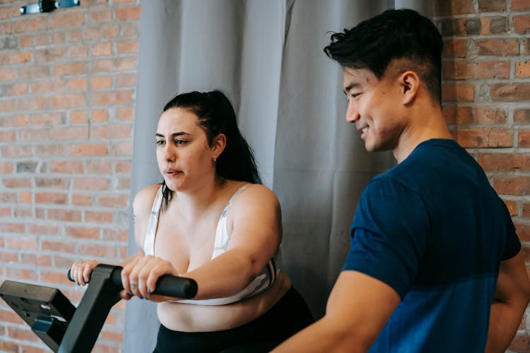 Smiling Young Ethnic Male Instructor Helping Obese Woman Training On Cycling Equipment